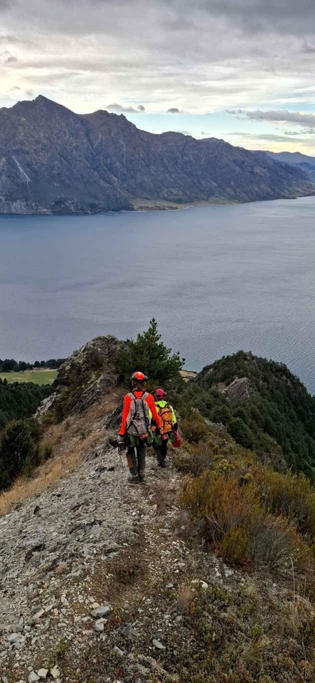 two people in work safety gear walking down a steep ridge on a mountain. A lake is below and mountains are in the background