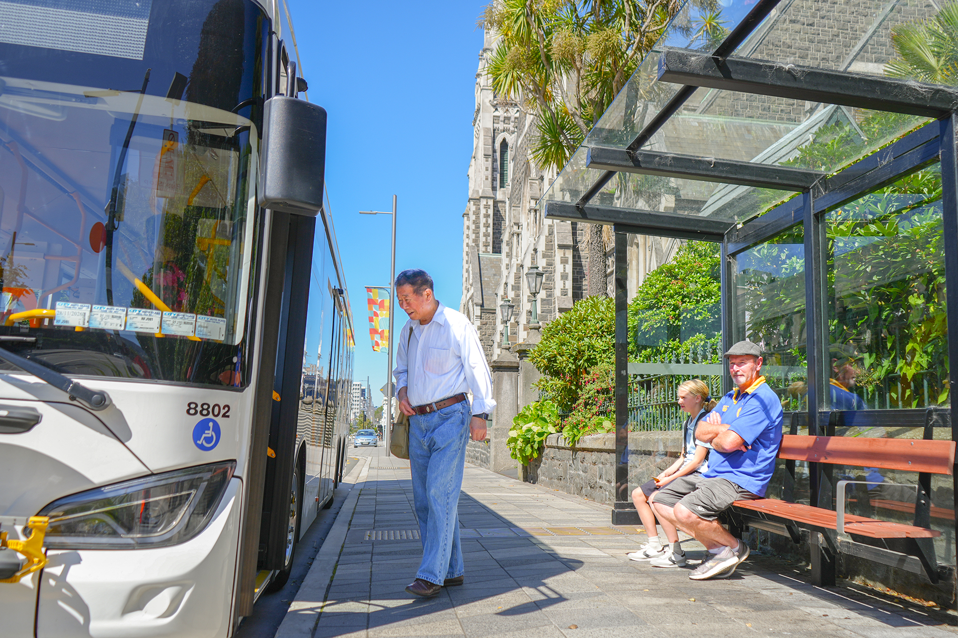 a person boarding a bus on a sunny day while two people sit at a bus stop