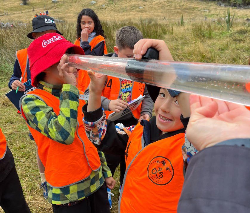 a group of children in orange shirts and hats holding up a plastic tube with water in it and looking through the bottom