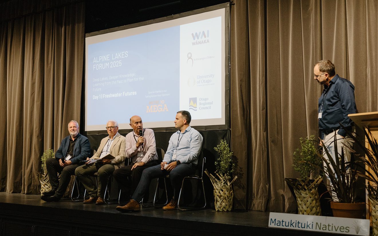 Four people sitting on chairs on a stage with a slideshow behind them, and a man standing beside them beside a lecturn