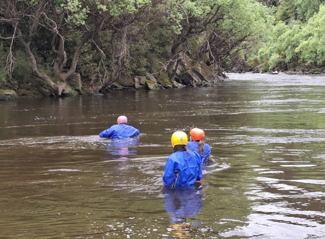 three people in waist-deep water wearing safety gear and crossing a river