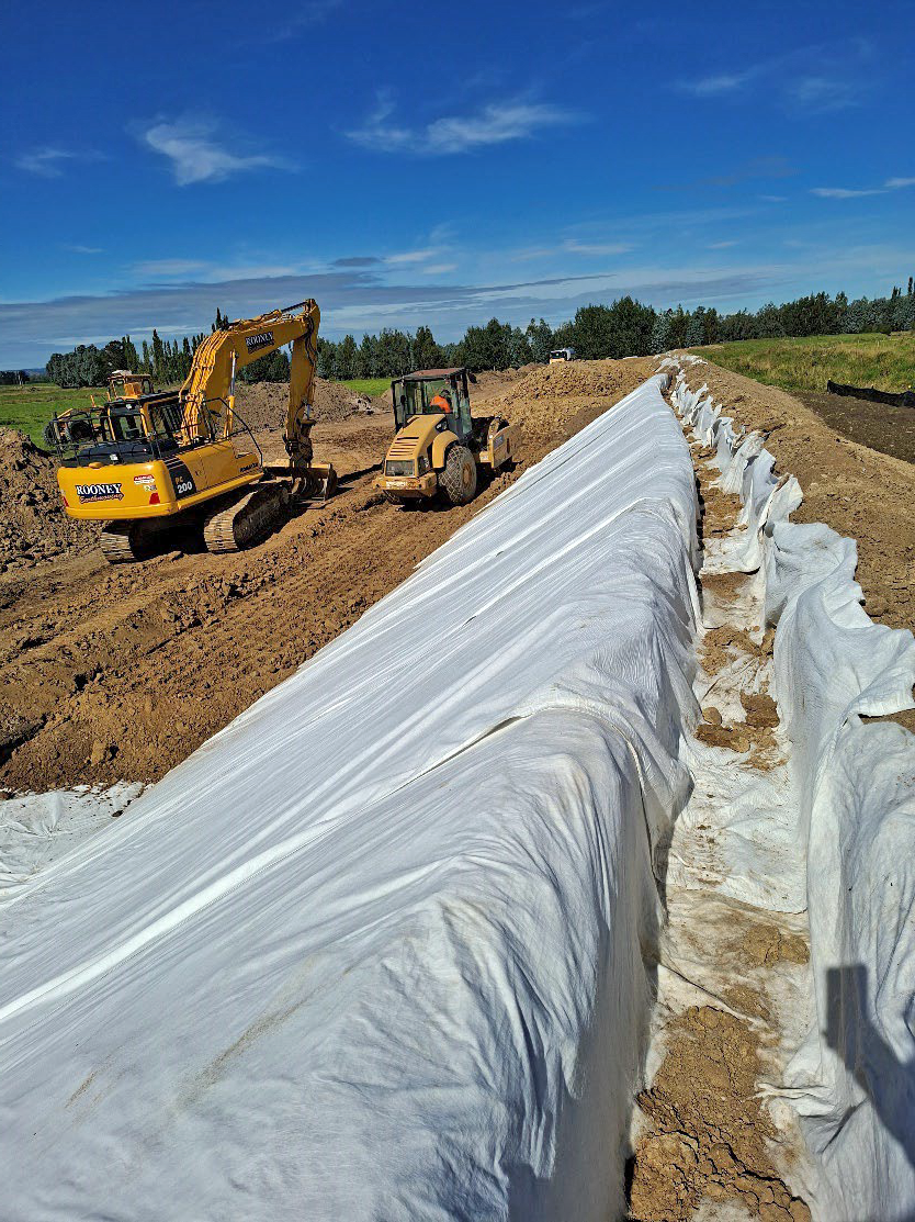 a digger and other earthworks machinery beside a long trench that has a white layer of fabric laid into it