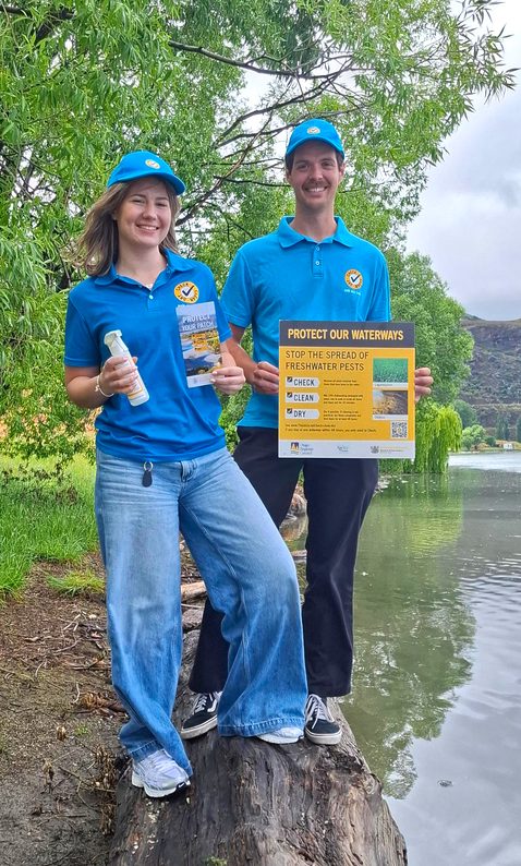 two people standing beside a lake holding a spray bottle and a sign and smiling at the camera