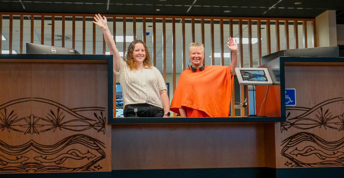 two smiling, waving people stand at a desk to welcome people