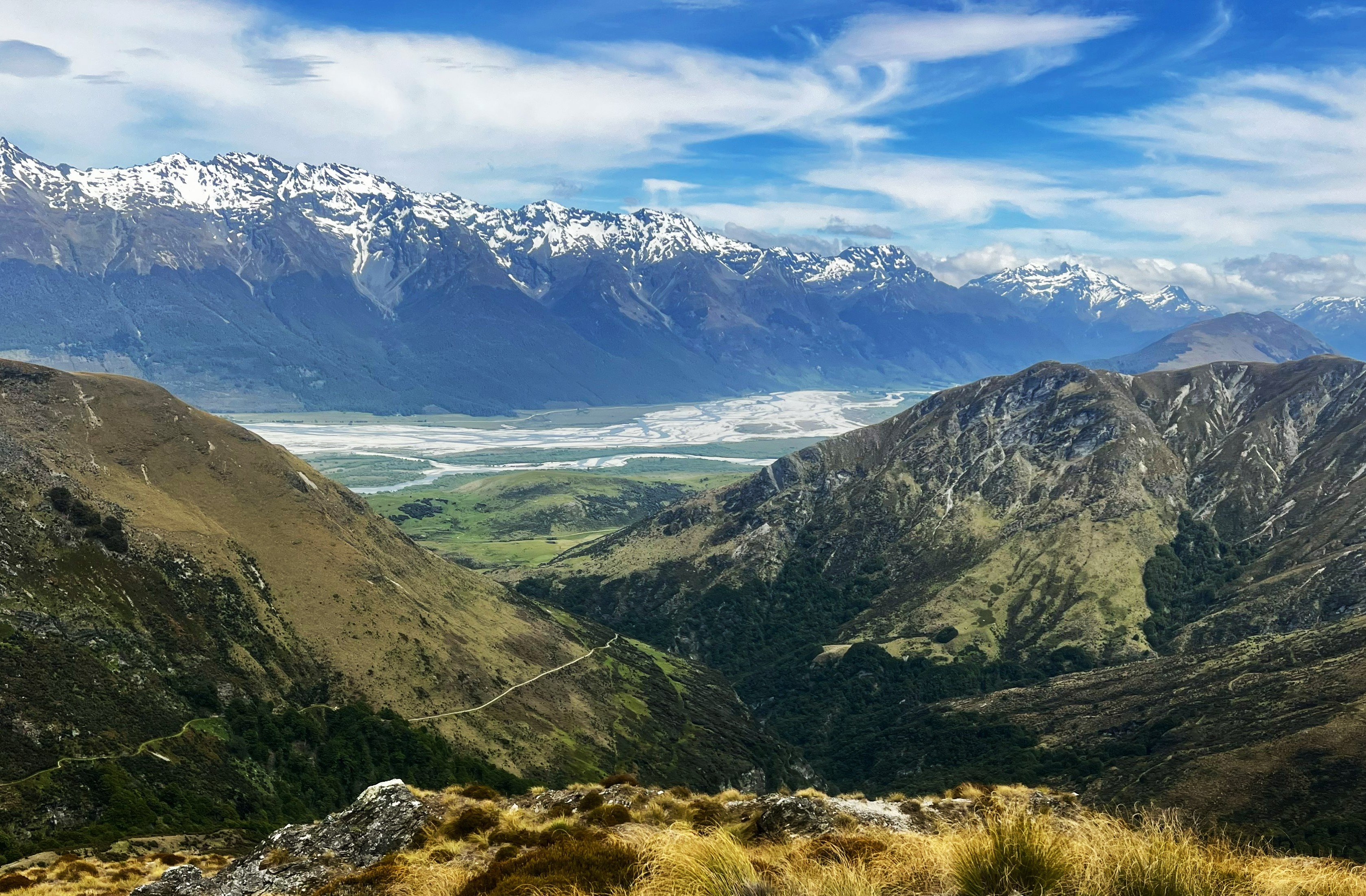 the view from the top of a mountain with a snow-capped mountain range in the distance and a river in the valley below