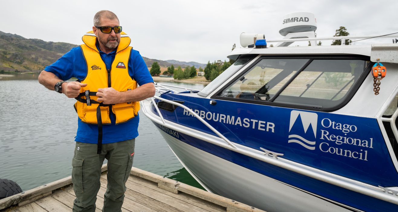 a man standing on a dock next to a boat putting on a lifejacket. The boat has with the words 'Harbourmaster' and 'Otago Regional Council' on it