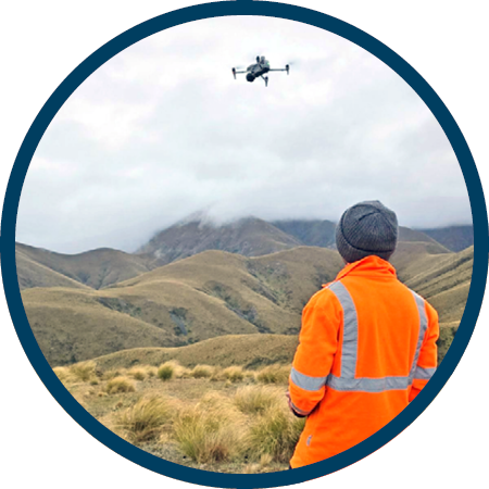 a person with their back to the camera wearing a high-visibility top and flying a drone over tussocky ground