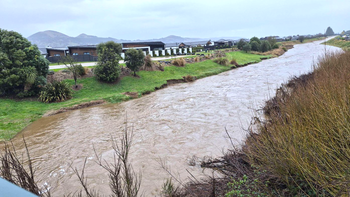 a stream with rushing brown water flowing through a semi rural area with houses nearby