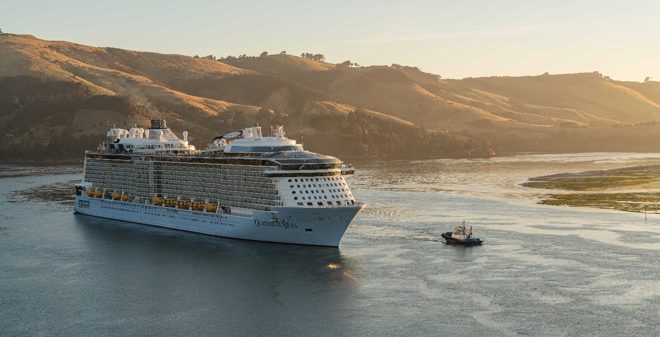 a large cruise ship in a harbour with a smaller tugboat nearby and hills in the background