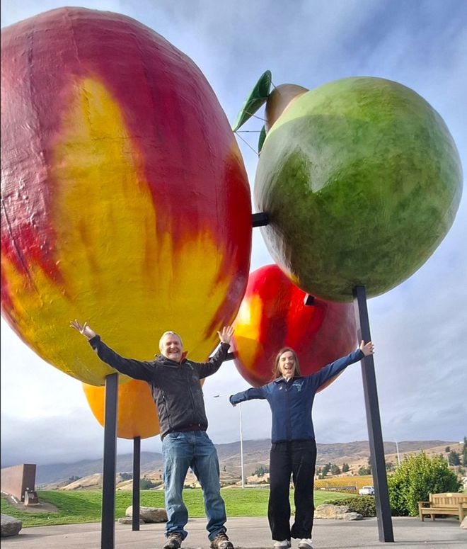 Two people with their arms in the air standing in front of four huge colourful fruit statues