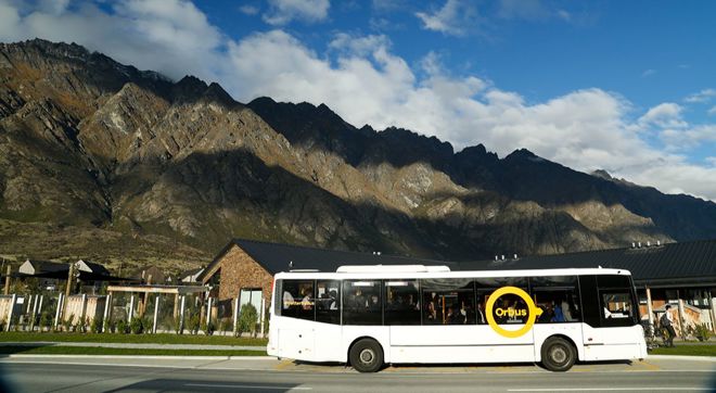 a bus dropping passengers at a stop with houses and mountains behind it