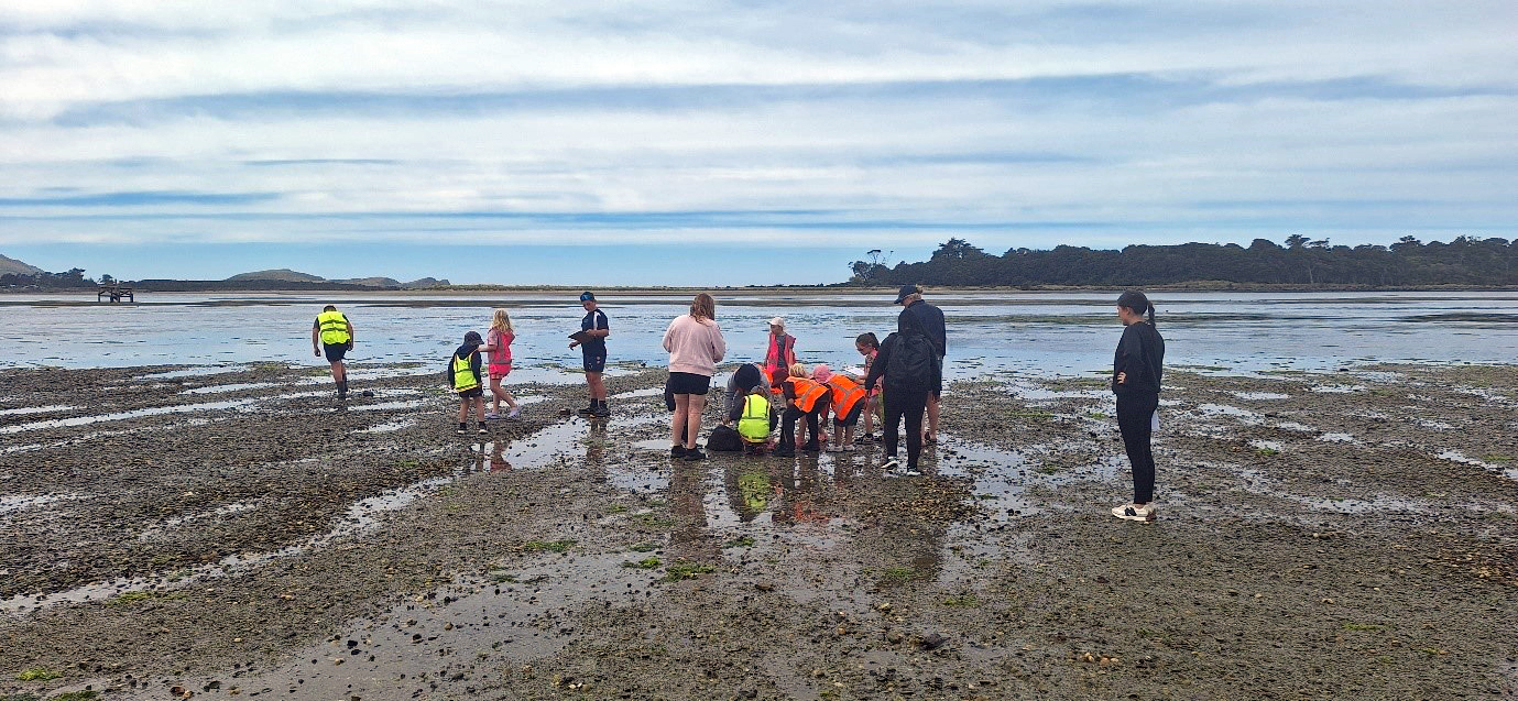 a group of children and some adults beside the sea. Some of the children are crouched down looking at the ground