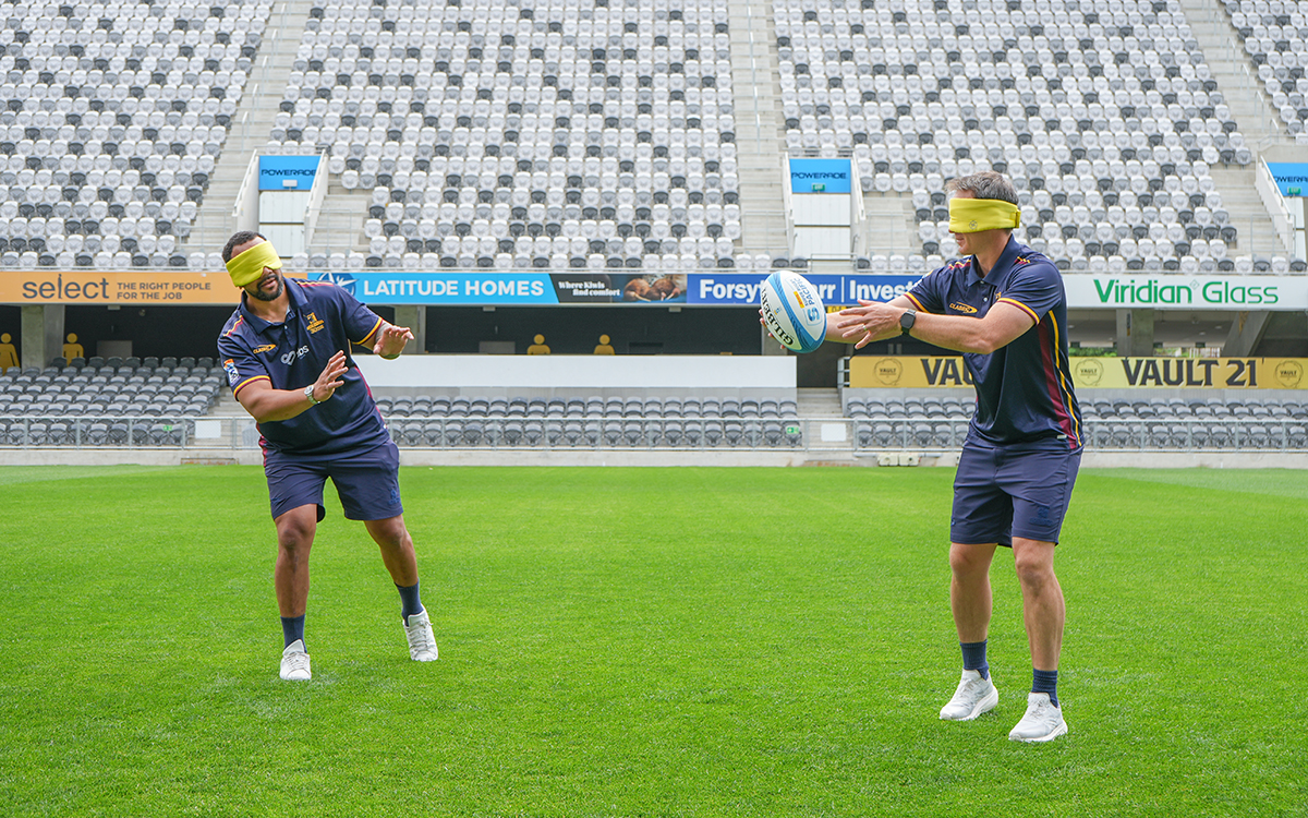 two rugby players in uniform with blinfolds on trying to throw a ball to each other on a field