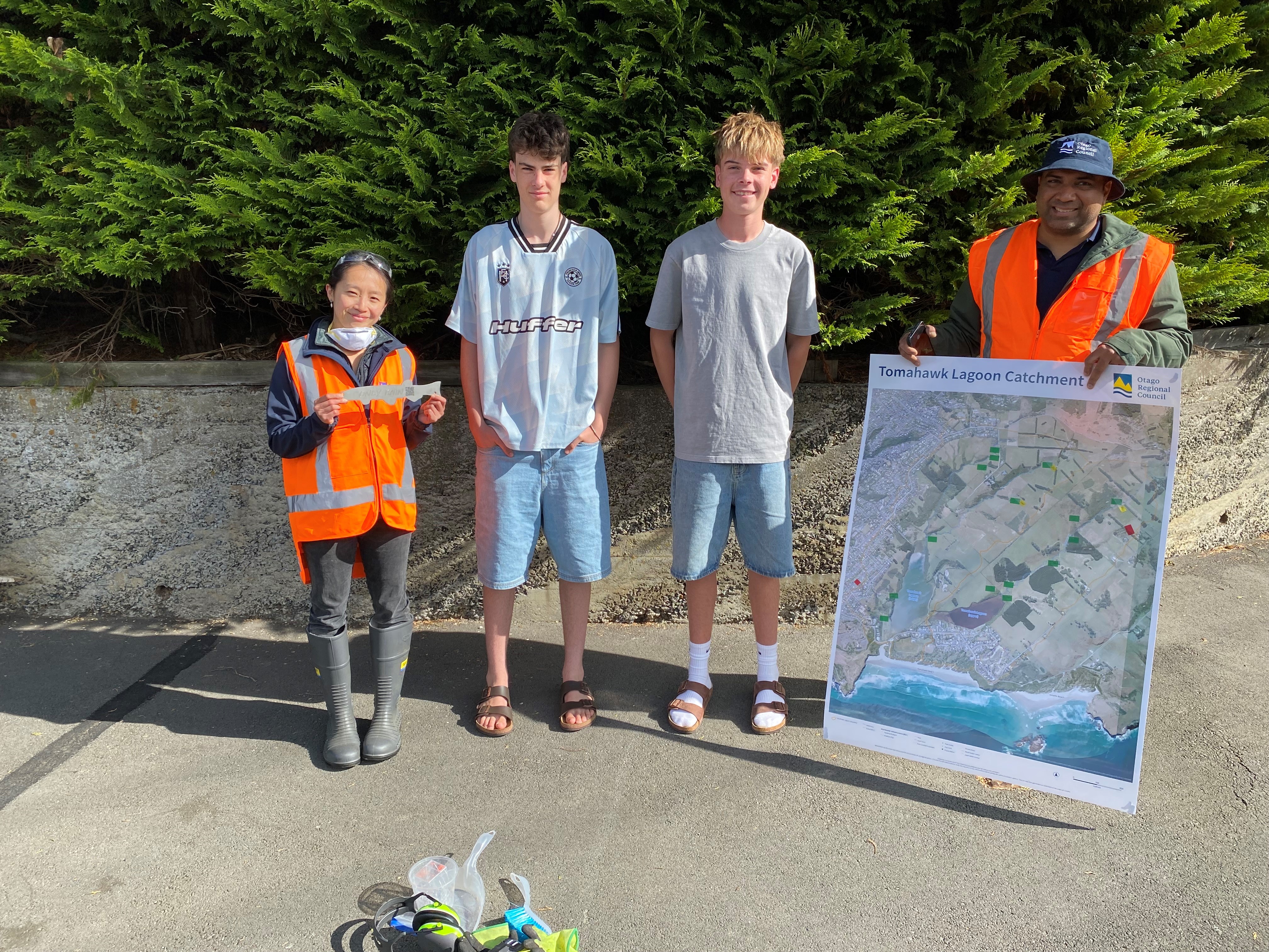 two Otago Regional Council staff with two teenagers. One of the staff memebers is holding up a metal fish and the other is holding a large poster showing the Tomahawk area