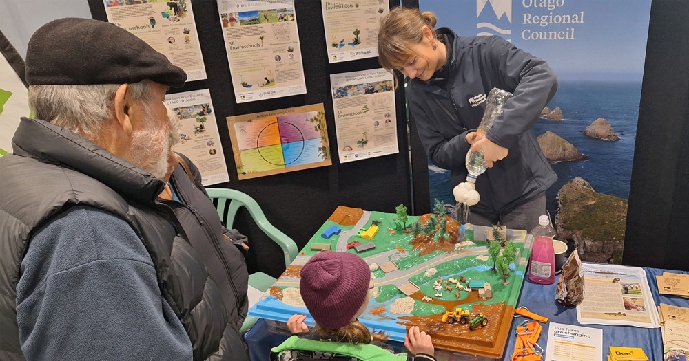 a person squirting water into a model of a landscape while a group of adults and a child watch