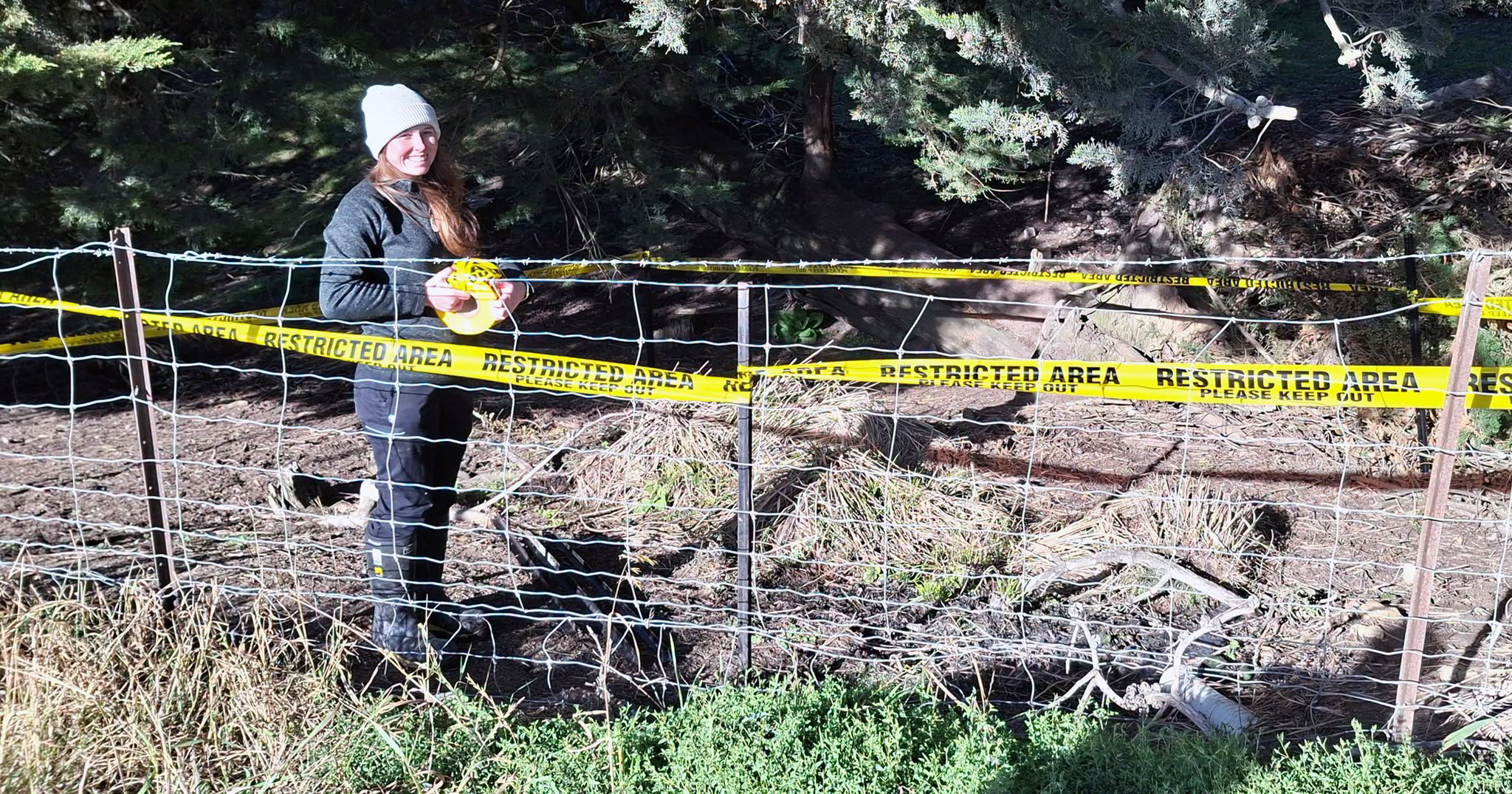 a person standing in front of a fence smiling at the camera and holding yellow tape with restricted area written on it