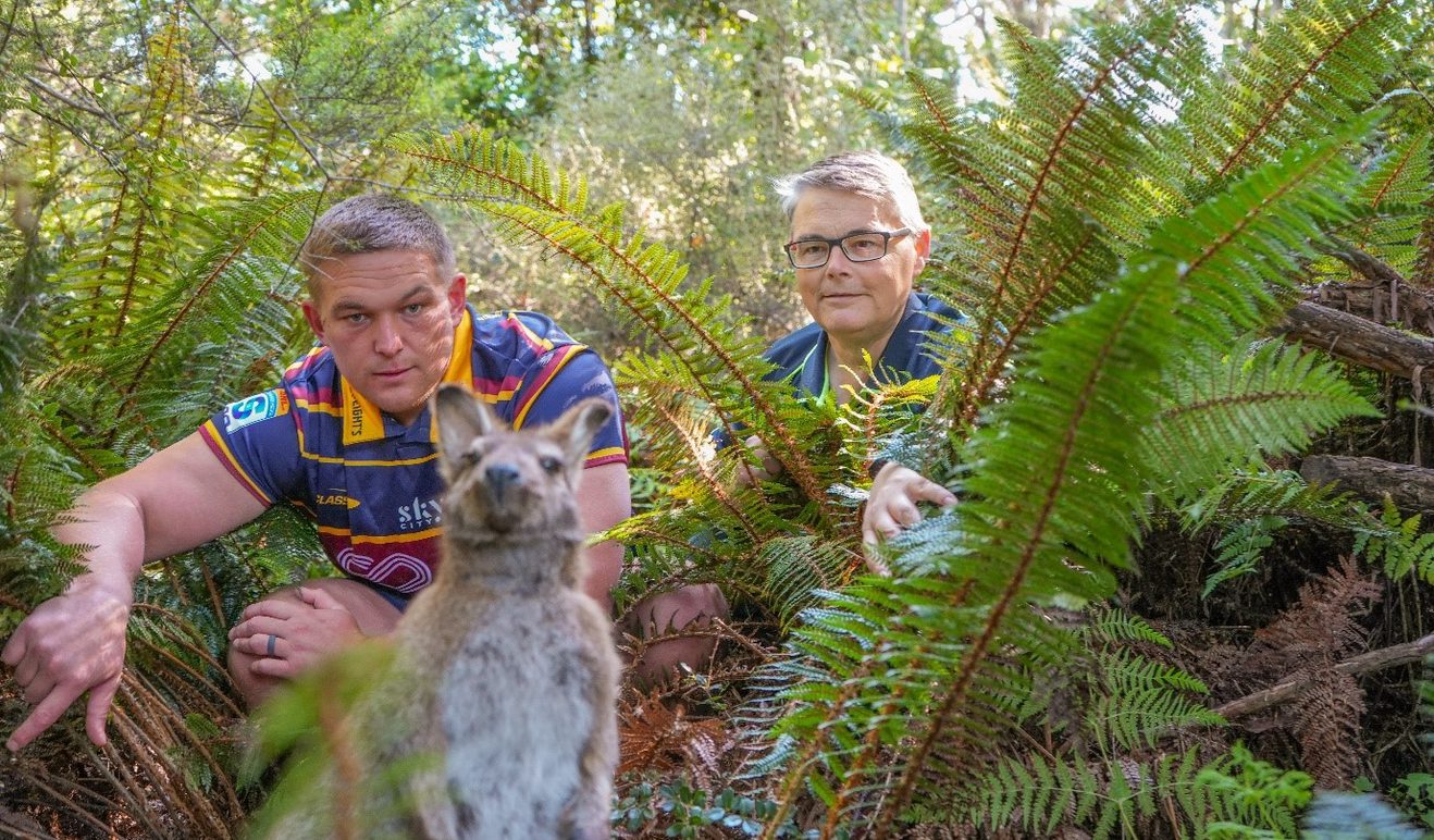 two people kneeling in the bush looking at a stuffed wallaby