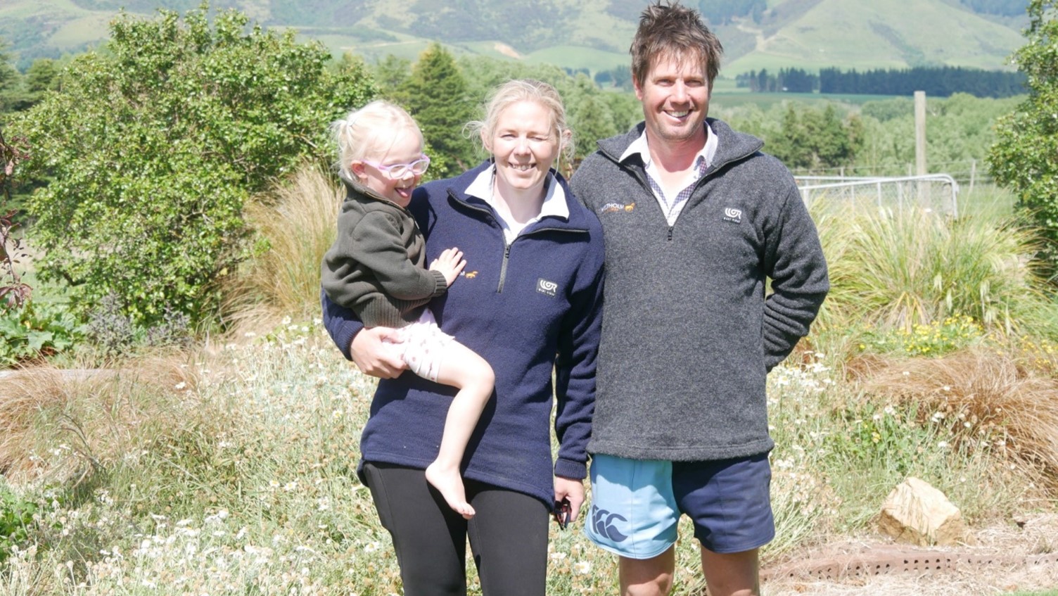 a couple in a rural setting smiling at the camera with one of them holding a small child
