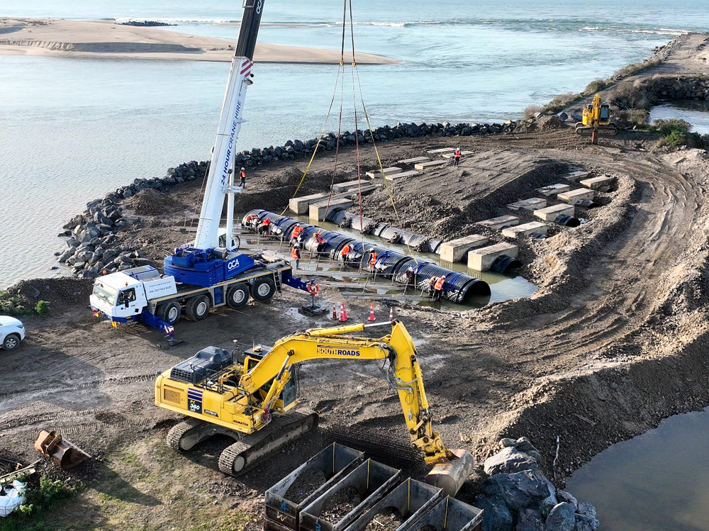 A construction site beside Puerua River with a crane and construction equipment. Workers in orange vests help to place a large black pipe into a trench.